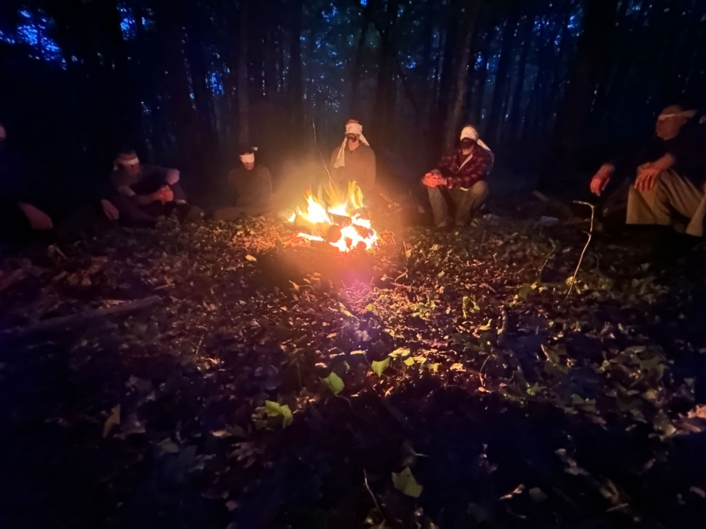 Cercle nocturne au feu, participants les yeux bandés — La Forge Groupe assis en cercle autour du feu de camp la nuit, yeux bandés, exercice de La Forge